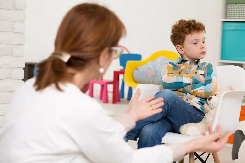 Woman therapist talking to the boy sitting on a chair with his arms folded