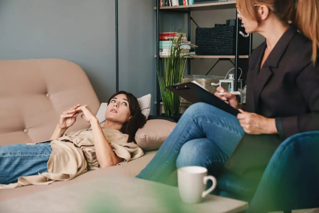 Photo of cute Caucasian woman lying on sofa and conversating with a psychologist on therapy session in room emphasizing the different types of mental health therapy