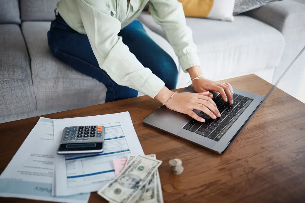 Computer, money and calculator with hands for home budget, financial planning and salary, rent cost or online loan. Person typing on laptop and sofa with cash and documents for mortgage and bills.