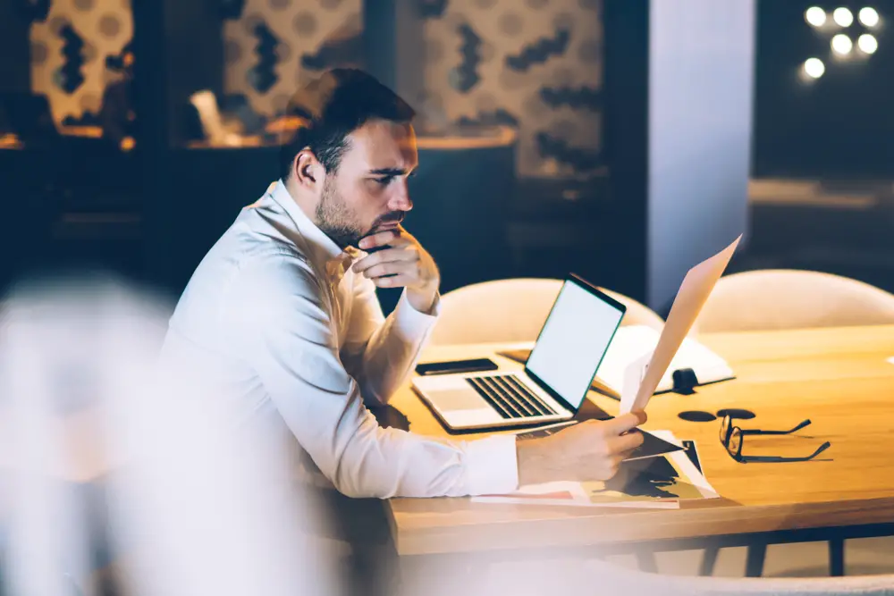 Side view of concentrated stubbled brunet entrepreneur holding document and touching chin with hand while working with laptop at table in meeting room