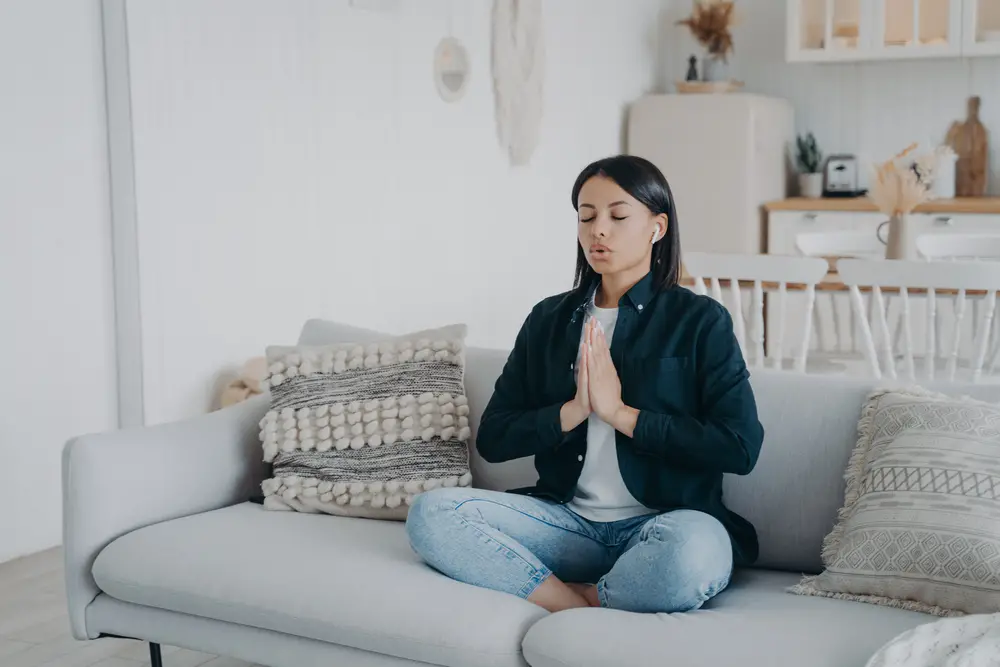 Calm young woman deep breathing practicing yoga sitting on couch at home. Female meditates folded hands in namaste gesture in lotus posture on sofa. Wellness, stress relief concept.